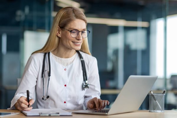 A medical professional wearing a stethoscope around her neck is using her laptop and writing something on her clipboard.