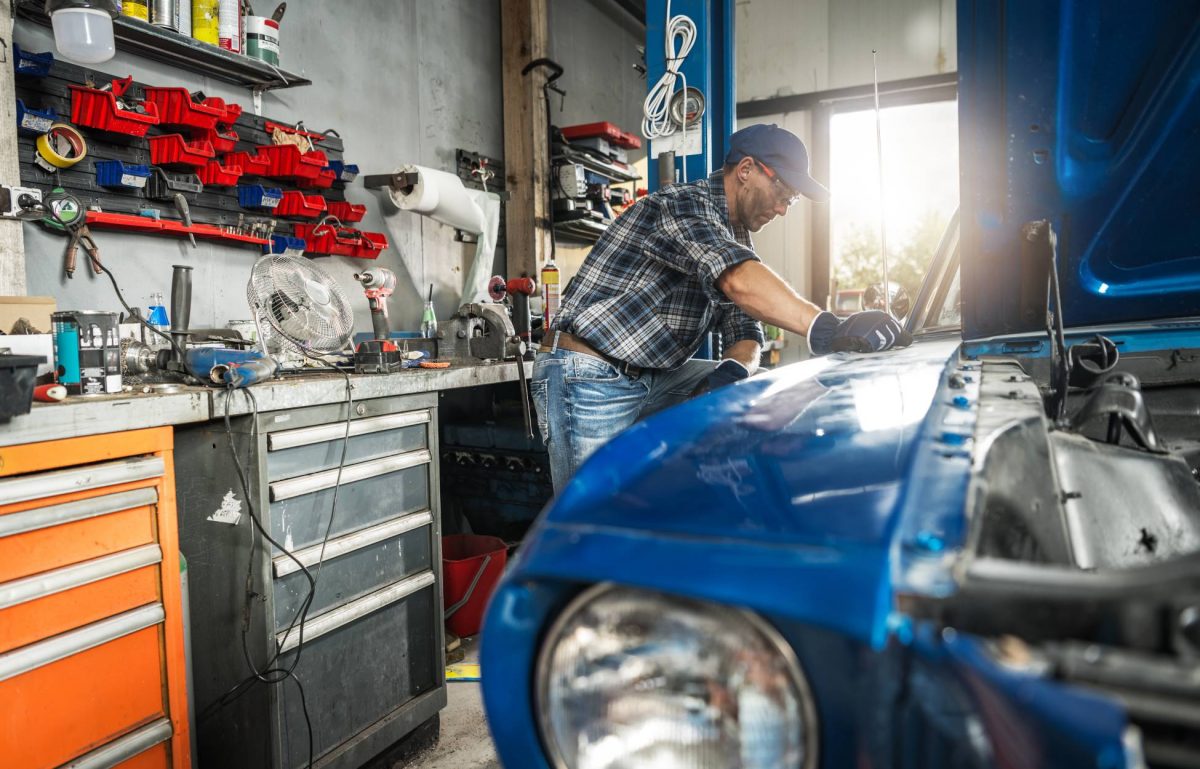 A man in a flannel, jeans, baseball hat, and safety glasses examines a blue, classic car with the hood up in a garage.