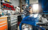 A man in a flannel, jeans, baseball hat, and safety glasses examines a blue, classic car with the hood up in a garage.