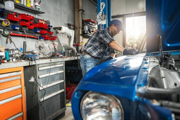 A man in a flannel, jeans, baseball hat, and safety glasses examines a blue, classic car with the hood up in a garage.