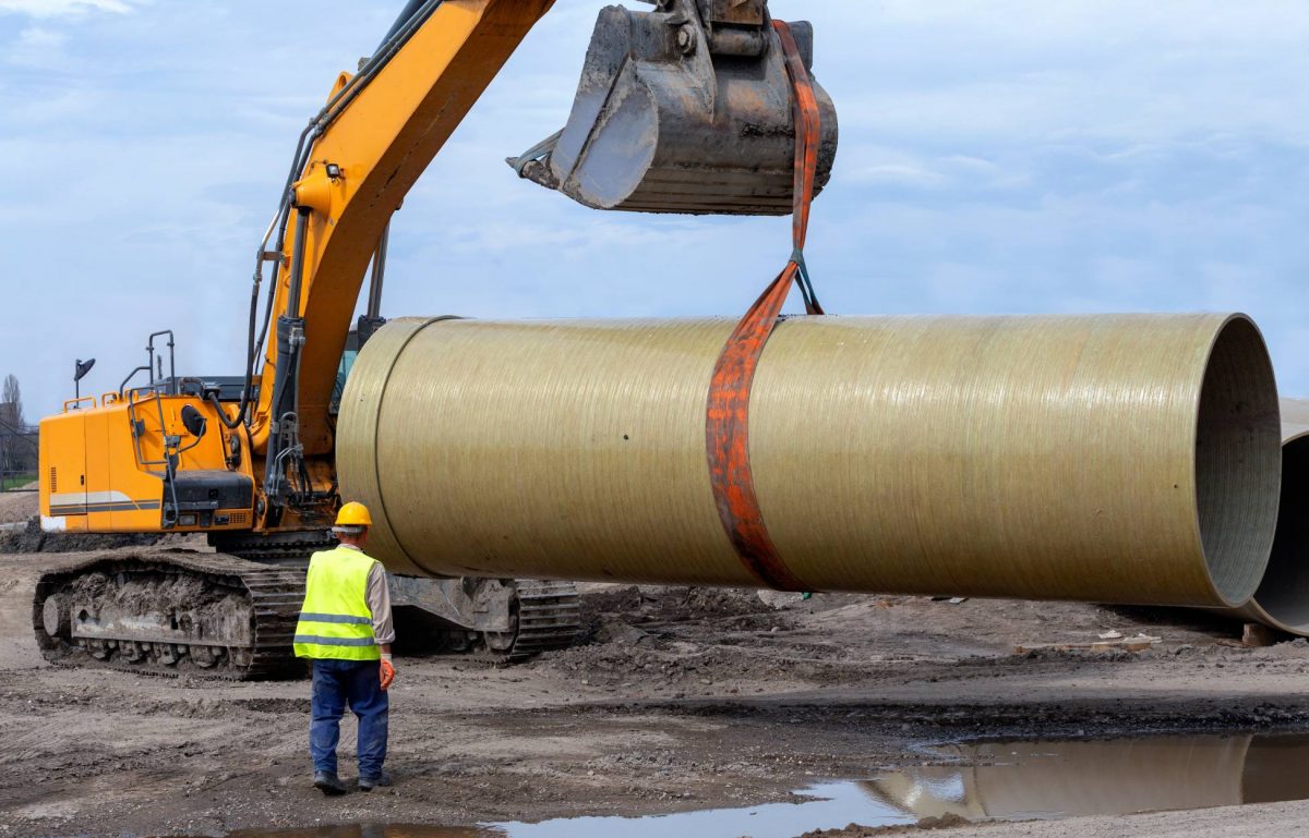 A worker is standing in front of an excavator with a bucket attachment, which is using an industrial band to lift a large pipe.