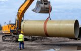 A worker is standing in front of an excavator with a bucket attachment, which is using an industrial band to lift a large pipe.