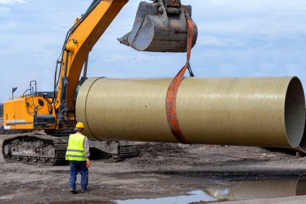 A worker is standing in front of an excavator with a bucket attachment, which is using an industrial band to lift a large pipe.