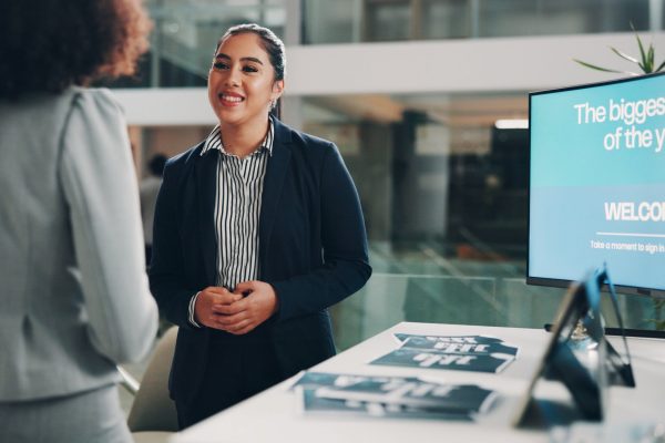 A smiling woman in business attire talking to another woman who's walked up to her convention booth.