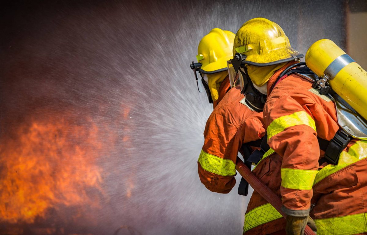 Two firefighters wearing orange and yellow uniforms face away from the camera, spraying water onto a fire.