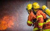 Two firefighters wearing orange and yellow uniforms face away from the camera, spraying water onto a fire.