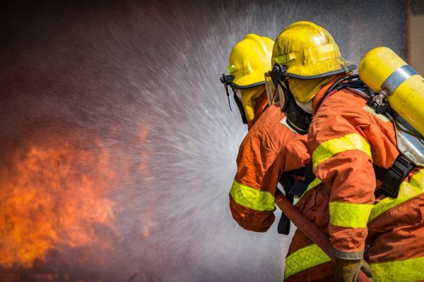 Two firefighters wearing orange and yellow uniforms face away from the camera, spraying water onto a fire.