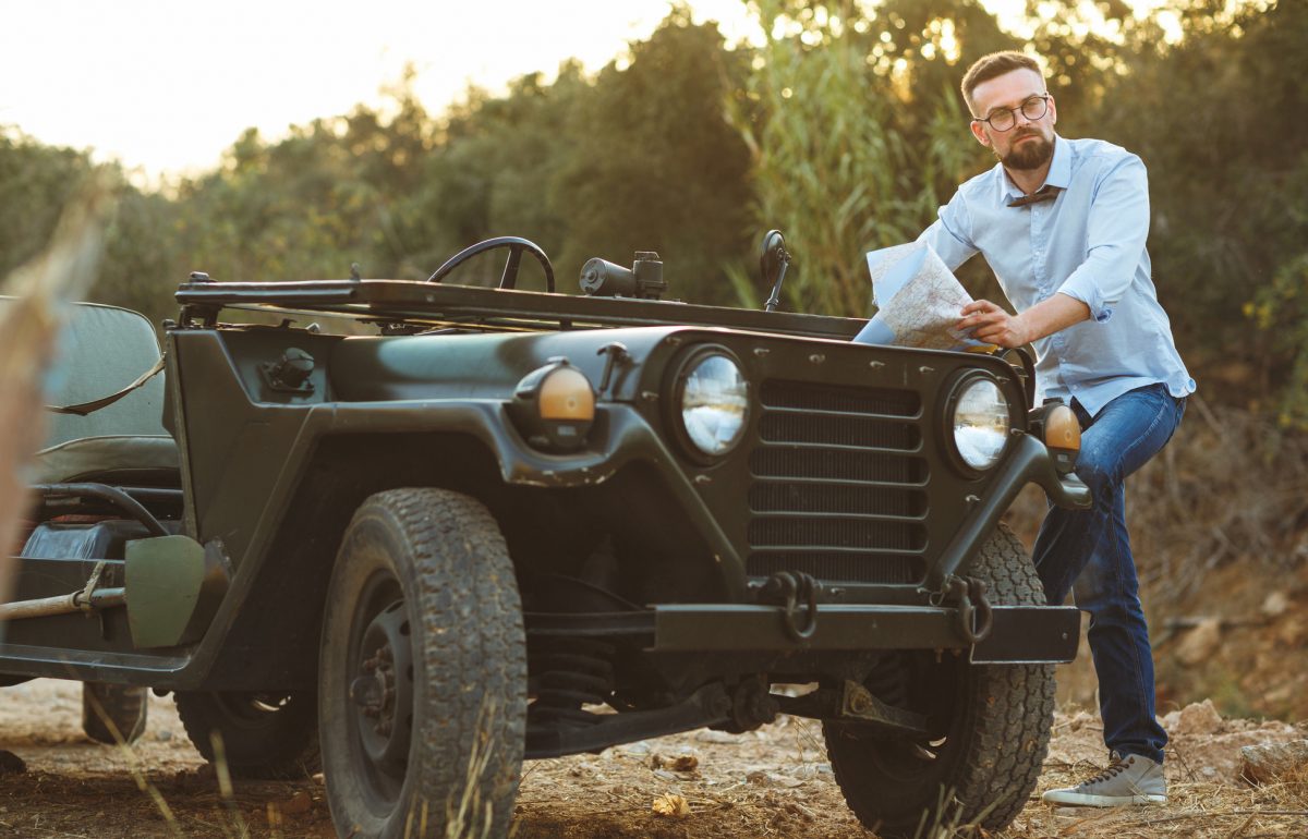 A man with a beard, bow tie, and black-rimmed glasses is resting his foot on the running board of a vintage Jeep.