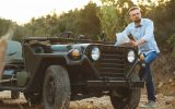 A man with a beard, bow tie, and black-rimmed glasses is resting his foot on the running board of a vintage Jeep.