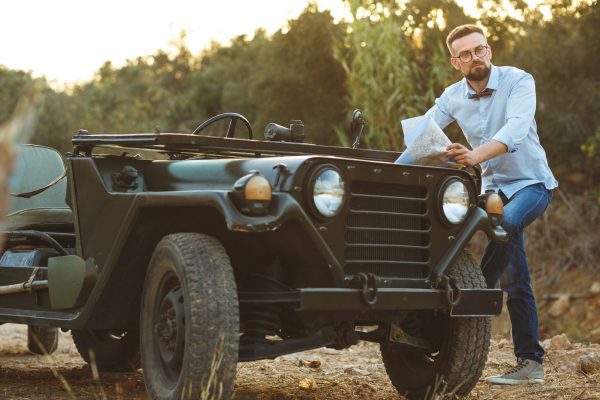 A man with a beard, bow tie, and black-rimmed glasses is resting his foot on the running board of a vintage Jeep.