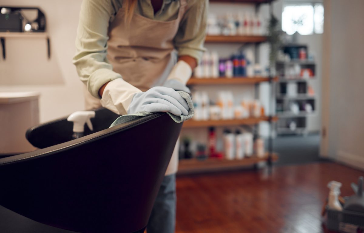 A woman cleaning the back of a styling chair in her salon. She's wearing gloves and using a cloth to wipe it down.