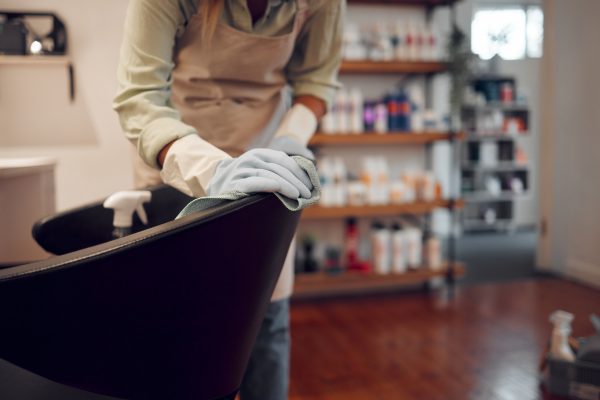 A woman cleaning the back of a styling chair in her salon. She's wearing gloves and using a cloth to wipe it down.