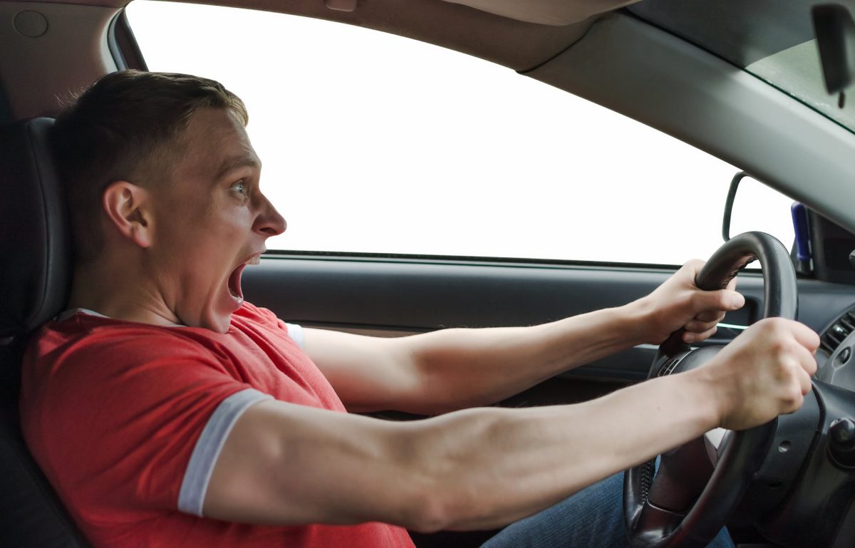 A man in a red shirt tightly gripping the steering wheel of his car and shouting as he seems to lose control of the vehicle.