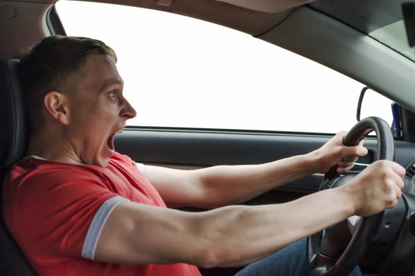 A man in a red shirt tightly gripping the steering wheel of his car and shouting as he seems to lose control of the vehicle.