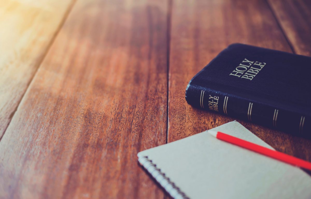 A wooden table with a Bible and a notepad and pencil. The pencil is bright red and the notepad is white.