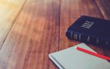 A wooden table with a Bible and a notepad and pencil. The pencil is bright red and the notepad is white.