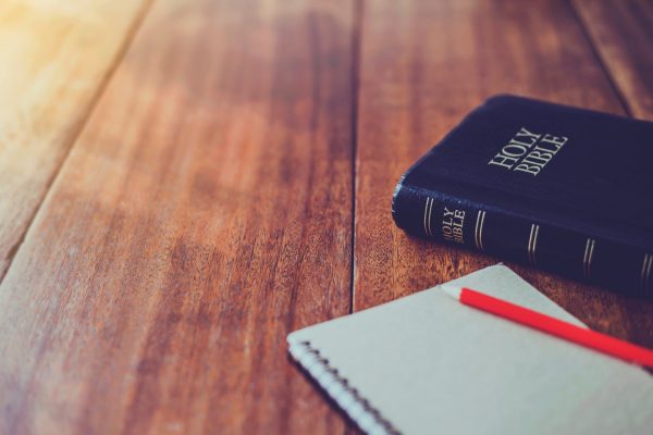 A wooden table with a Bible and a notepad and pencil. The pencil is bright red and the notepad is white.