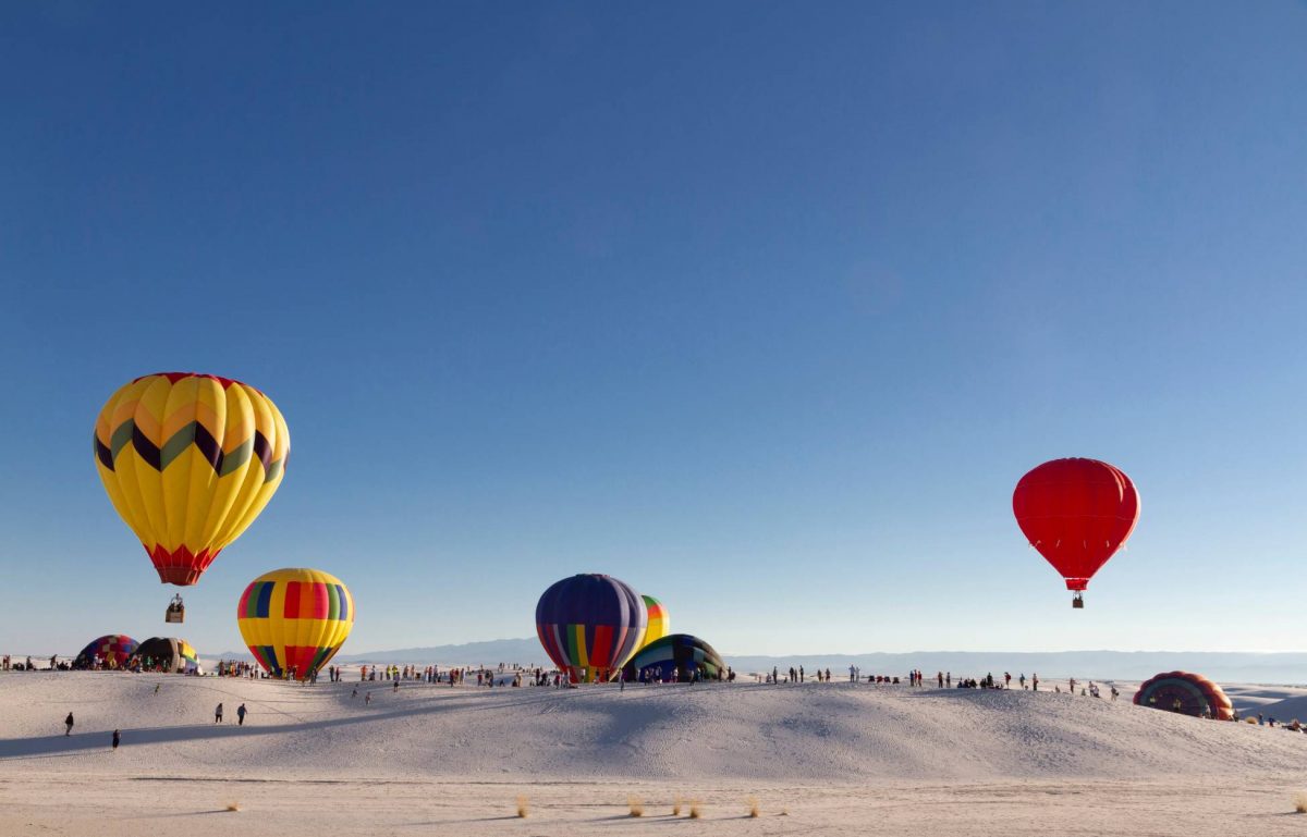 Several large and colorful hot air balloons hovering above White Sands National Park, framed against the sand and blue sky.