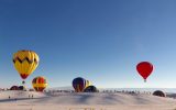 Several large and colorful hot air balloons hovering above White Sands National Park, framed against the sand and blue sky.