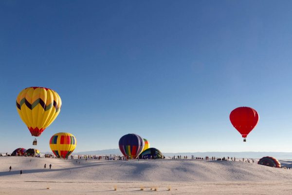 Several large and colorful hot air balloons hovering above White Sands National Park, framed against the sand and blue sky.