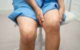 A person sitting on a white stool wearing blue garments. Their knees have vertical scars from knee replacement surgery.