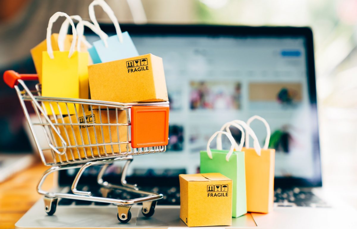 A toy shopping cart with miniature boxes and shopping bags sits on a laptop. The laptop has a blurred product page.