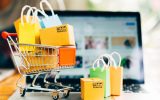 A toy shopping cart with miniature boxes and shopping bags sits on a laptop. The laptop has a blurred product page.