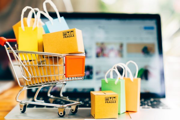 A toy shopping cart with miniature boxes and shopping bags sits on a laptop. The laptop has a blurred product page.