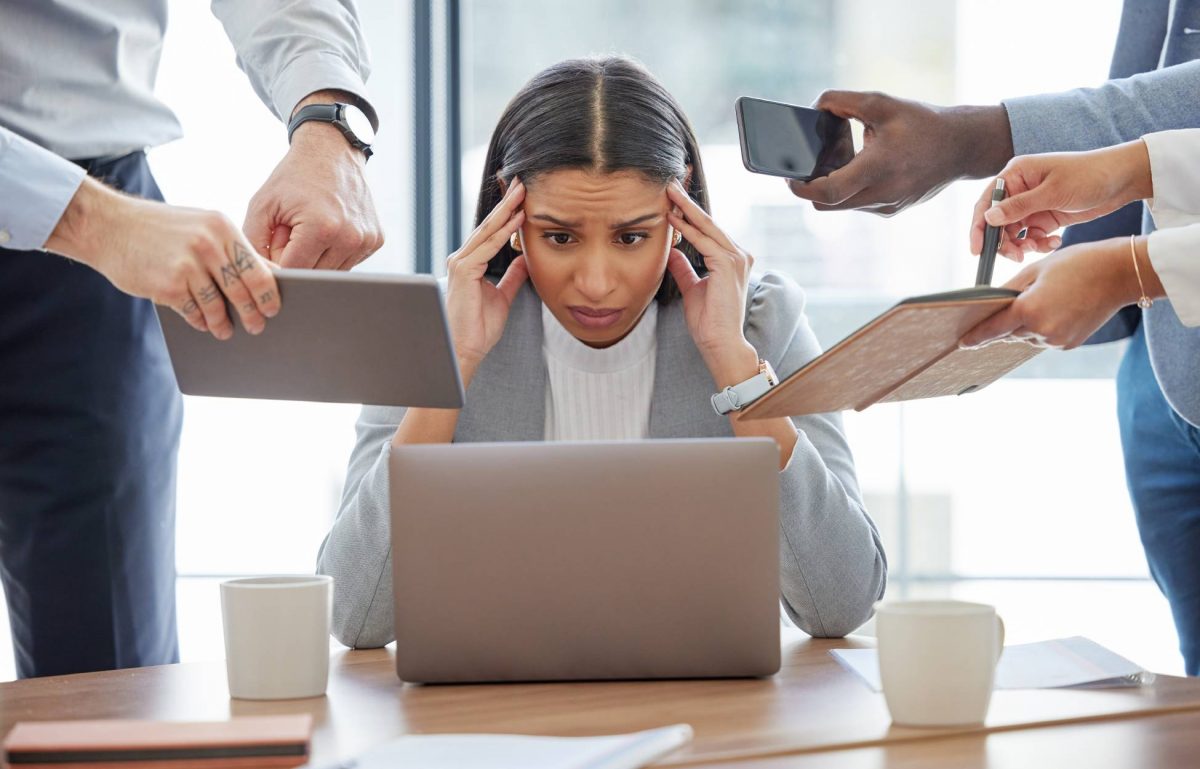 A woman sits at a desk looking at a laptop with her hands on her head and a distressed look on her face. People are by her.