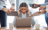 A woman sits at a desk looking at a laptop with her hands on her head and a distressed look on her face. People are by her.