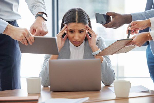 A woman sits at a desk looking at a laptop with her hands on her head and a distressed look on her face. People are by her.