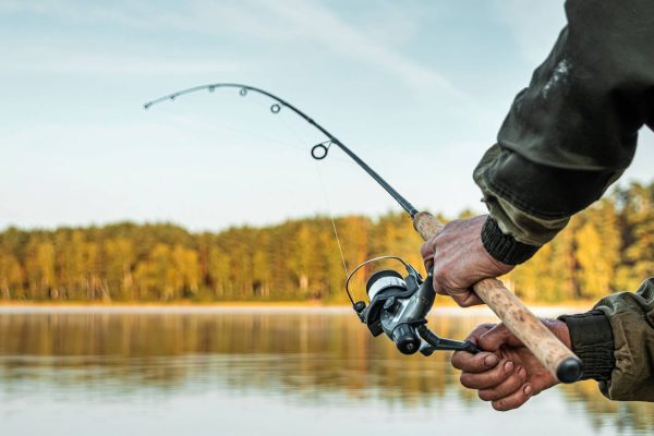 A close-up view shows a person with a dark-colored coat holding a wooden handle fishing pole. Trees are in the background.