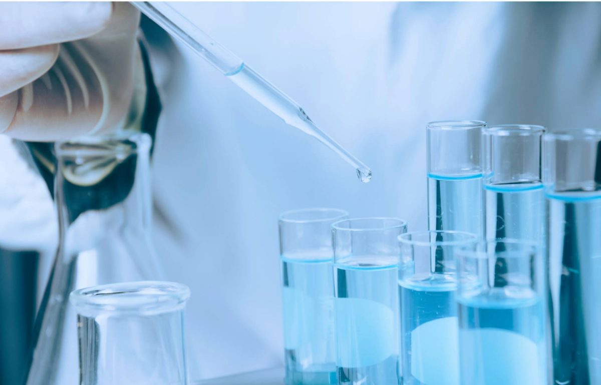 Close-up to multiple sample tubes filled with a clear blue liquid while a scientist pours drops into one with a dropper.