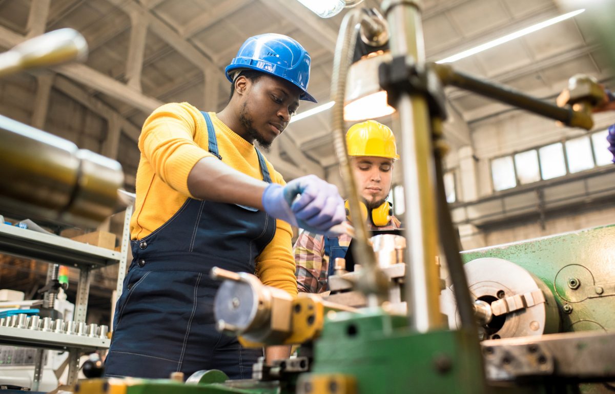 Two workers wearing blue and yellow uniforms working on a large metal machine inside a warehouse environment.