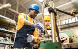 Two workers wearing blue and yellow uniforms working on a large metal machine inside a warehouse environment.