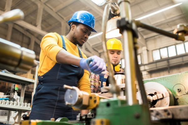 Two workers wearing blue and yellow uniforms working on a large metal machine inside a warehouse environment.