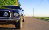 A close-up of a black-and-white vintage Mustang car. The vehicle is parked on the side of the road in a rural area.