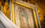 A large painting of a woman in a light pink dress and a teal shawl hangs on a golden wall. The woman is praying.