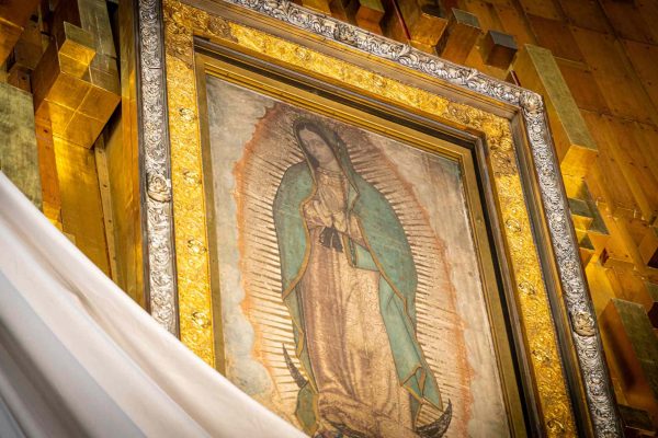 A large painting of a woman in a light pink dress and a teal shawl hangs on a golden wall. The woman is praying.