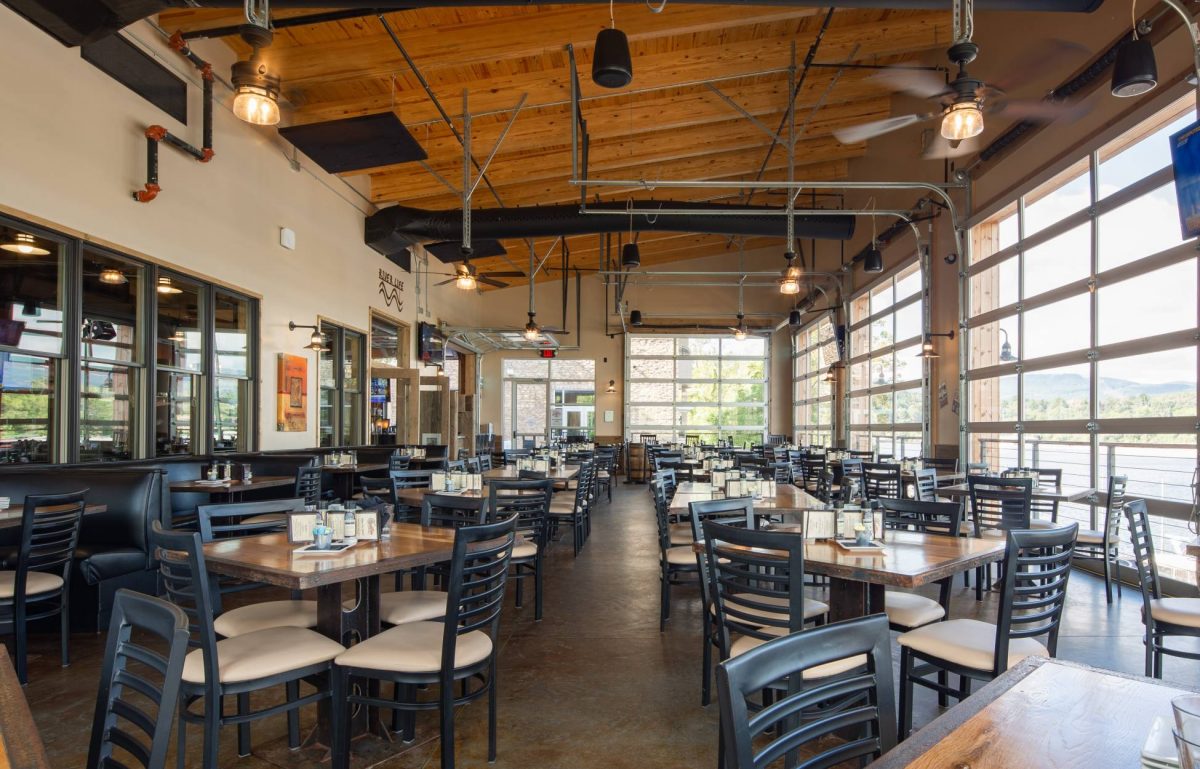 A riverside restaurant patio with black chairs and small wooden tables, enclosed with glass restaurant garage doors.