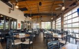 A riverside restaurant patio with black chairs and small wooden tables, enclosed with glass restaurant garage doors.