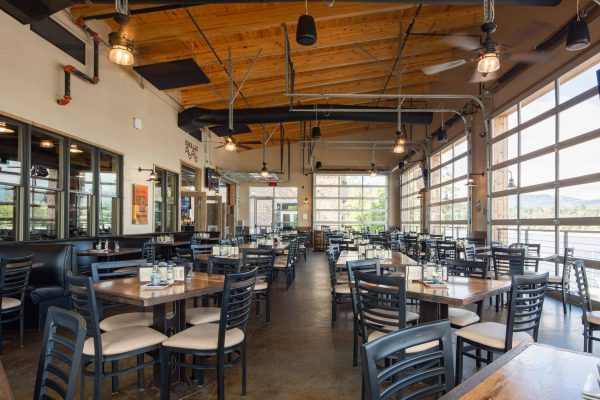 A riverside restaurant patio with black chairs and small wooden tables, enclosed with glass restaurant garage doors.