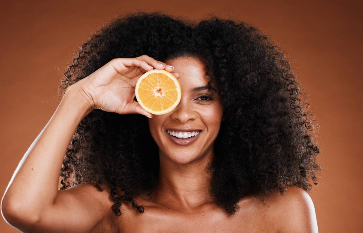 A Black woman with tight curly hair smiling with her teeth and holding an orange over her right eye.