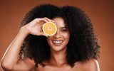 A Black woman with tight curly hair smiling with her teeth and holding an orange over her right eye.