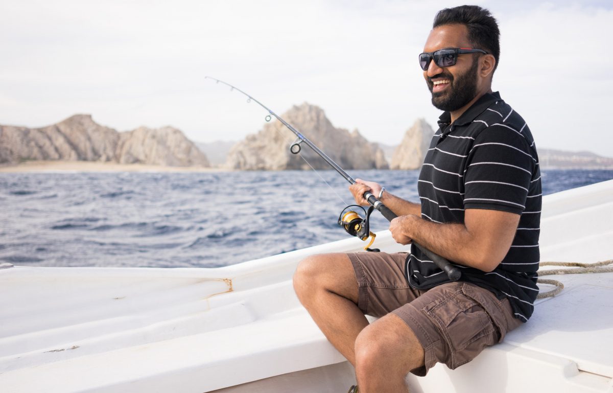 A young grinning man in cargo shorts and a striped tee, sitting on the edge of a boat and holding a fishing rod cast into the sea.