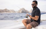 A young grinning man in cargo shorts and a striped tee, sitting on the edge of a boat and holding a fishing rod cast into the sea.