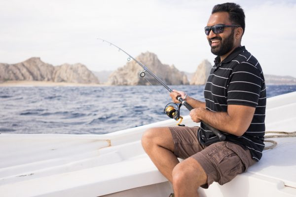 A young grinning man in cargo shorts and a striped tee, sitting on the edge of a boat and holding a fishing rod cast into the sea.