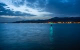 A body of water at night with city lights in the distance. In the middle of the water is a floating buoy with a green light.