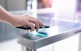 A person's hand holding a black smartphone over a green scanner built into a metal gate structure for access control.