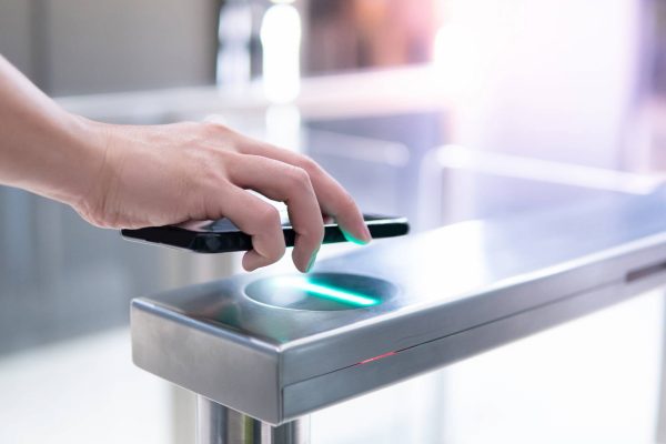 A person's hand holding a black smartphone over a green scanner built into a metal gate structure for access control.